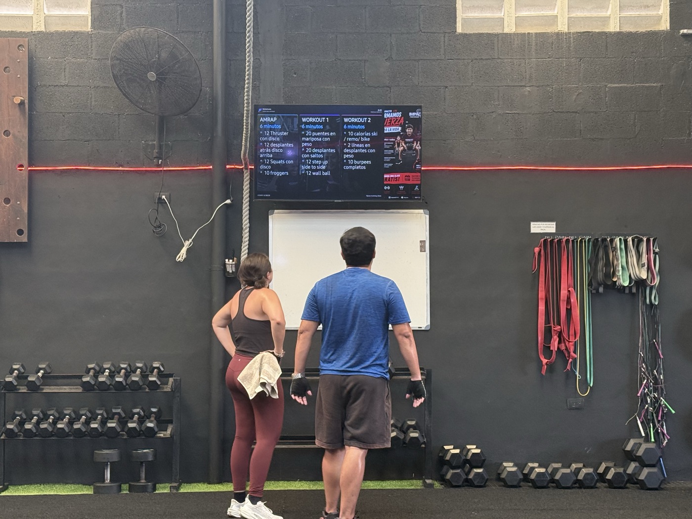 Socios consultando ejercicios y un anuncio en la pantalla del gimnasio
